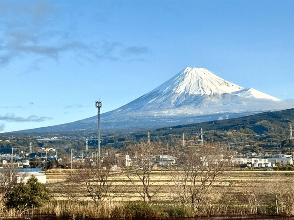 【富士山新幹線】青空の下で雪をまとった富士山が雄大に広がり手前には田畑と建物電波塔や鉄道橋が見える冬の景色