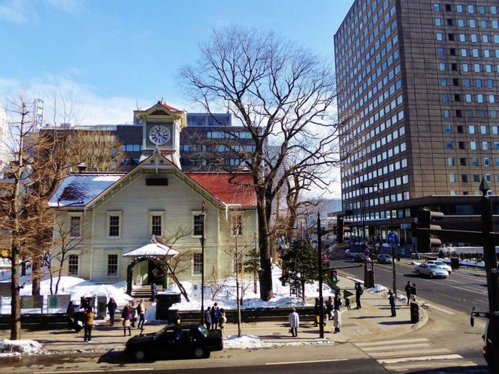 【日本三大がっかり名所】札幌時計台の白い建物に雪が積もり青空の下で人々が行き交う冬の風景
