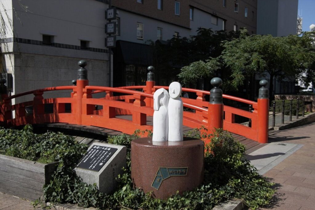 【日本三大がっかり名所】鮮やかな朱色の丸い形の橋の手前に白い二人の人型像と黒い石碑がある風景周りには緑の植物が生い茂る