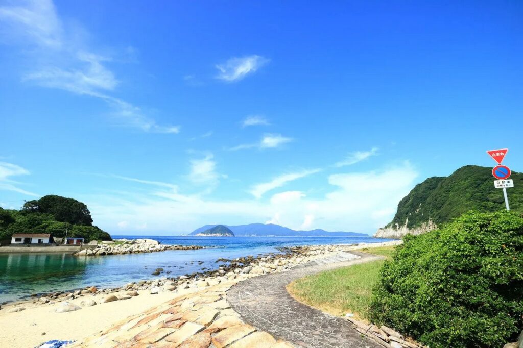 【柏島 がっかり】晴れた青空と透き通る海が広がる柏島の海岸線砂浜と岩場の間に小道が続く右手には緑の山々遠くには島々が見える