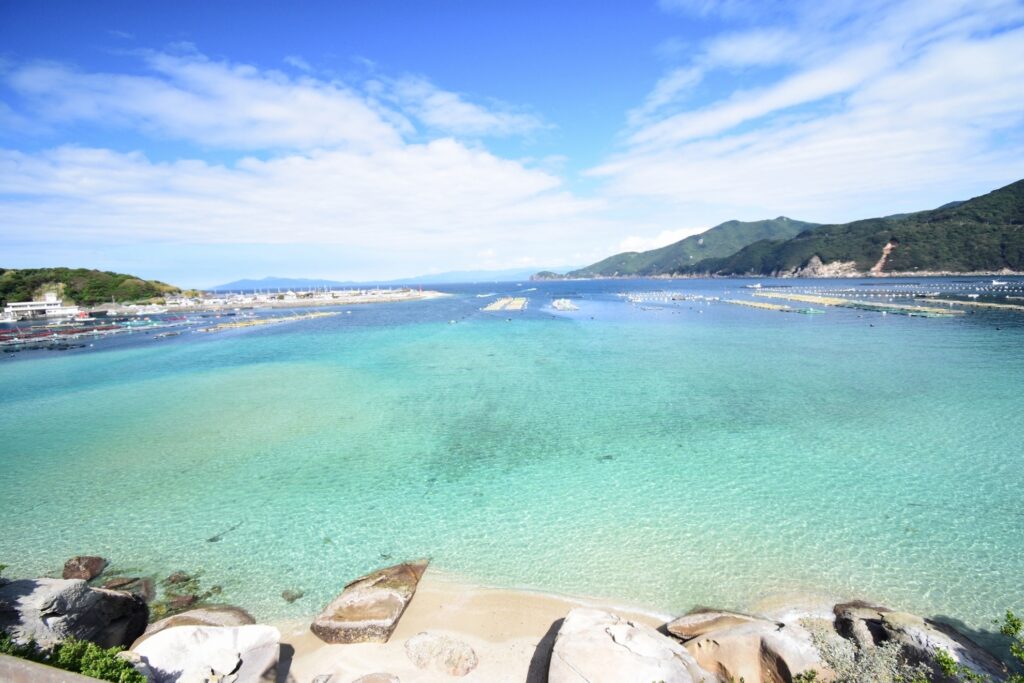 【柏島 がっかり】透き通ったエメラルドグリーンの海と白い砂浜 青空に白い雲が広がり遠くには海に浮かぶ養殖いかだと緑の山々が見える
