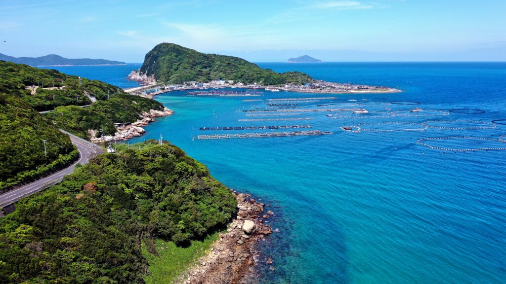 【柏島 がっかり】高知県柏島の透き通った青い海に魚の養殖いかだや丸い網が浮かぶ空撮写真 緑の山肌に沿ってカーブする道が見え奥には集落と漁港そして小さな島々が広がる風景