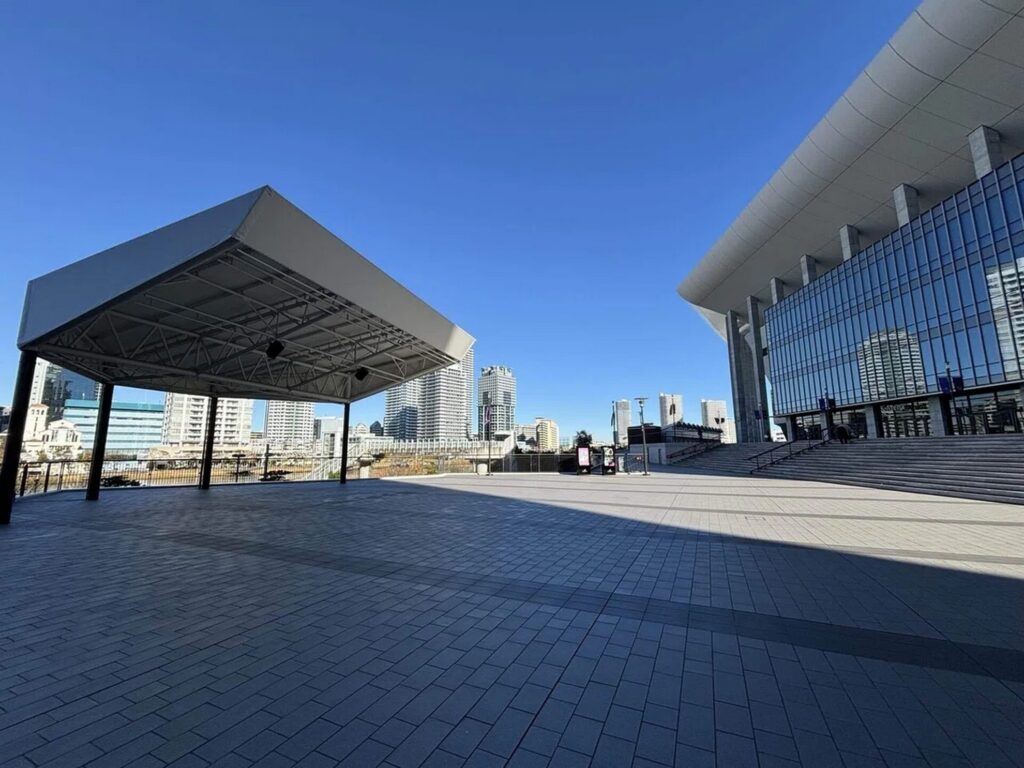 【歩道橋 ロケ地】晴れた青空の下に広がる石畳の広場と屋根付きステージ大きなガラス張りの建物と奥に見える高層ビル群