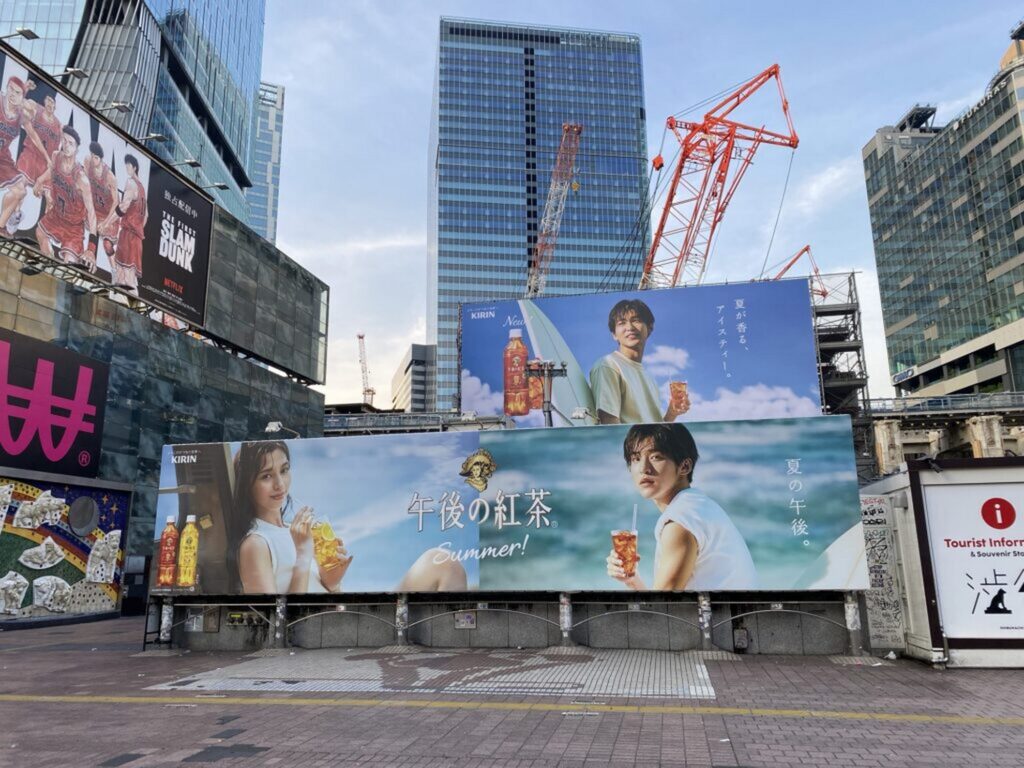 【すのちゅーぶ ロケ地】高層ビルと工事用クレーンのある街中にスラムダンクの看板と若い男女が写る午後の紅茶の大きな広告が並ぶ風景
