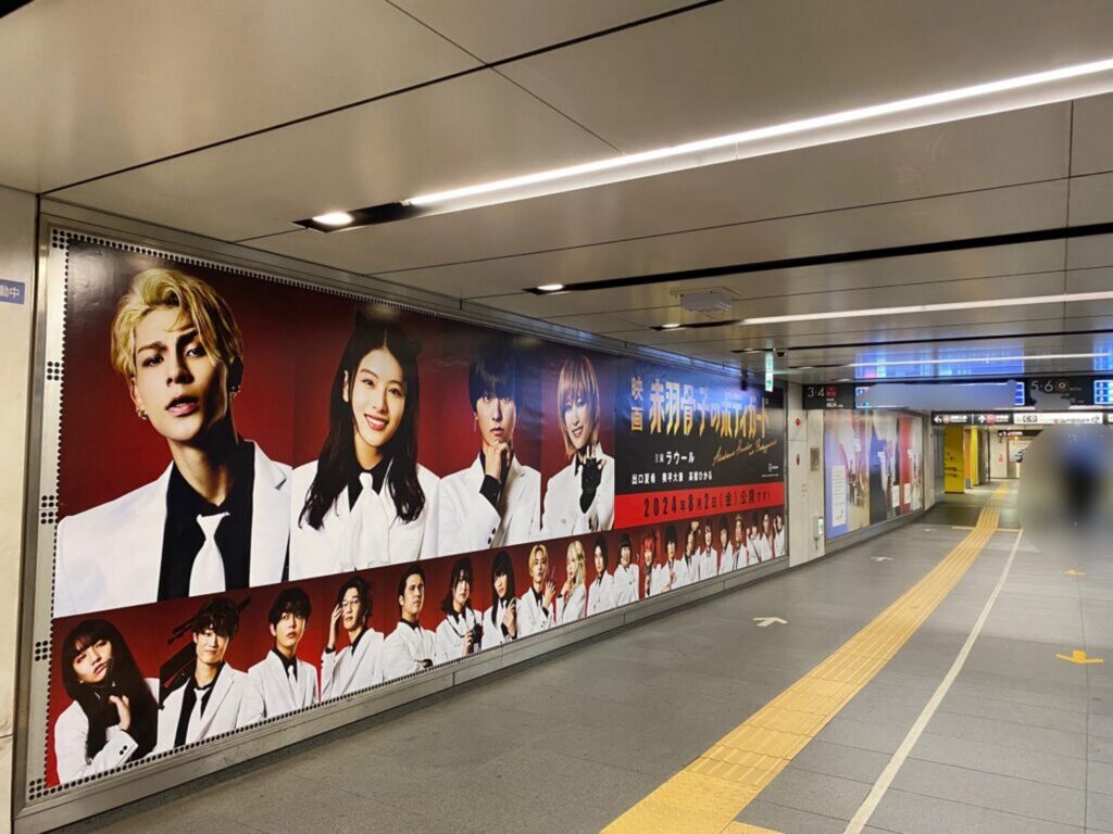 【すのちゅーぶ ロケ地】地下鉄の通路に掲示された赤い背景の大きな映画ポスター白と黒の衣装を着た複数の登場人物と公開日が書かれている