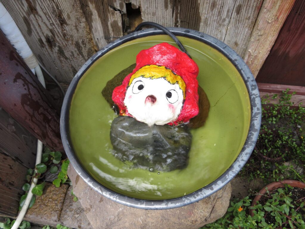 【鞆の浦がっかり】緑色に変色した水が溜まった丸い容器に赤い帽子と白い顔の人形が浮かんでいる 古い木の壁と植物