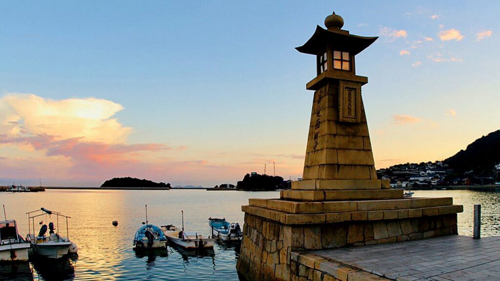 【鞆の浦がっかり】夕焼け空が広がる鞆の浦の港にそびえる常夜灯と水面に浮かぶ複数の小舟