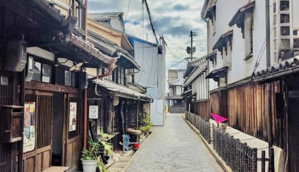 【鞆の浦がっかり】古く狭い路地に瓦屋根の木造家屋が並び生活感あふれる風景奥には工事中の白い覆いが見える曇り空