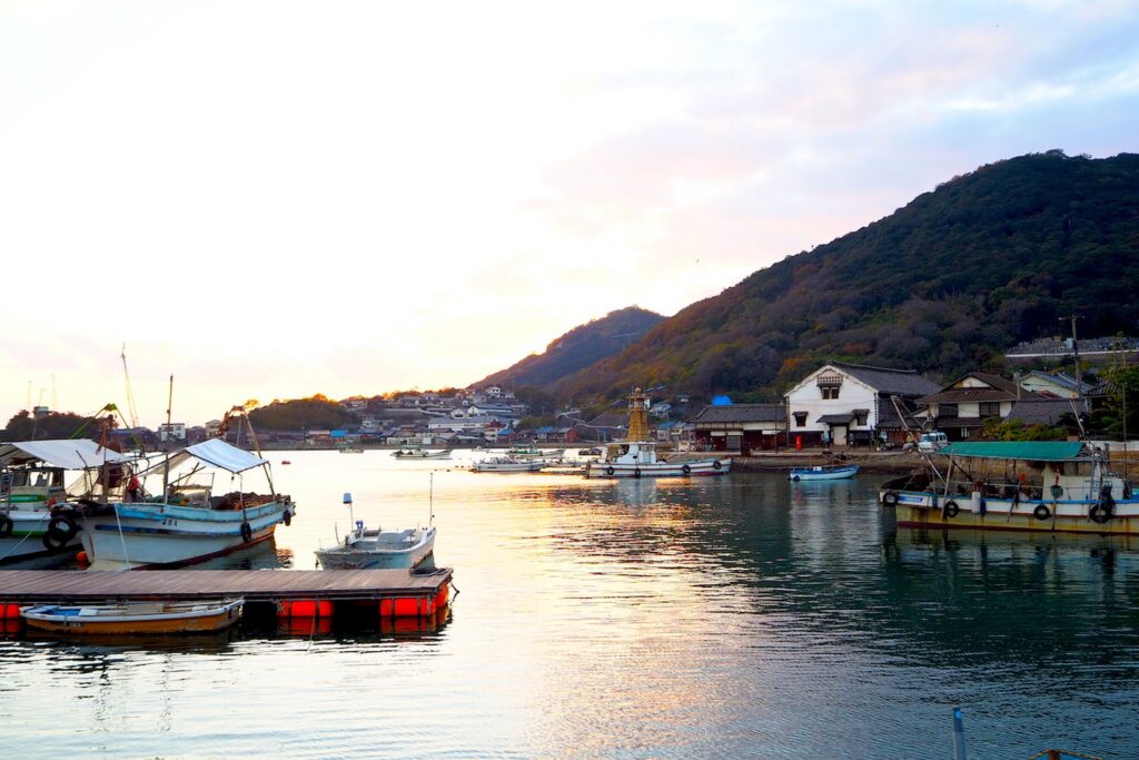 【鞆の浦がっかり】夕焼けに染まる港に停泊する漁船が並び山を背景に家々が立ち並ぶ