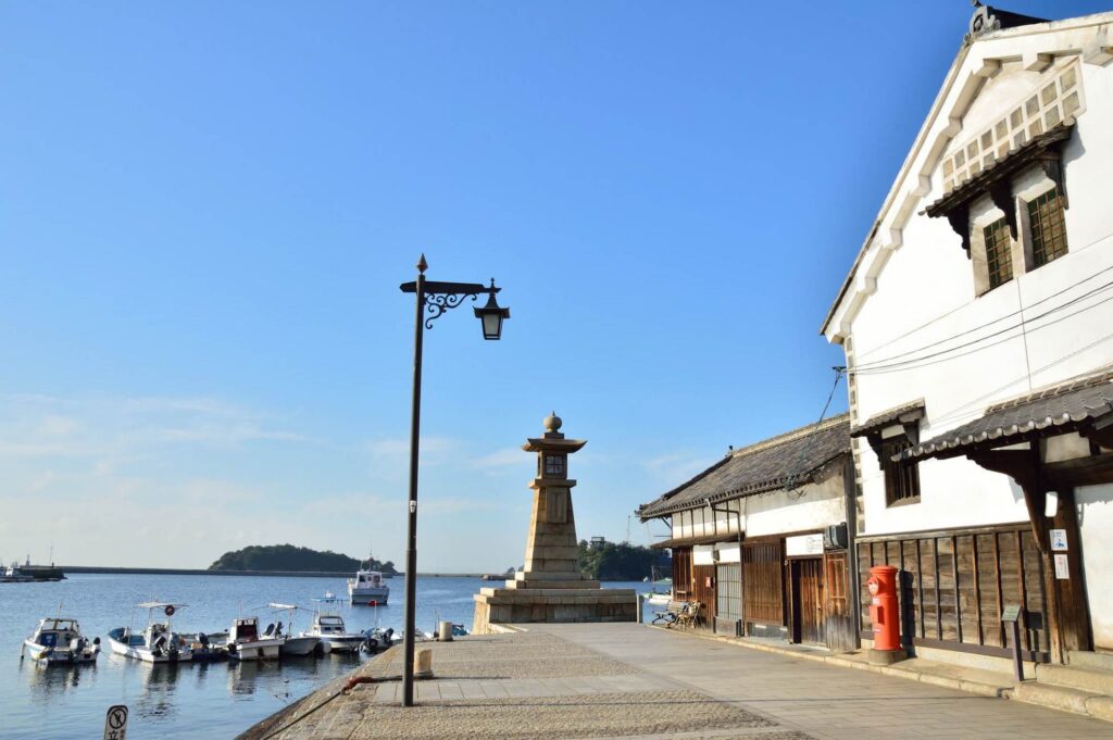 【鞆の浦がっかり】青い空の下海沿いに建つ常夜灯と白い壁の古い町並み停泊する多くの船と赤い郵便ポストがある日本の港町の風景