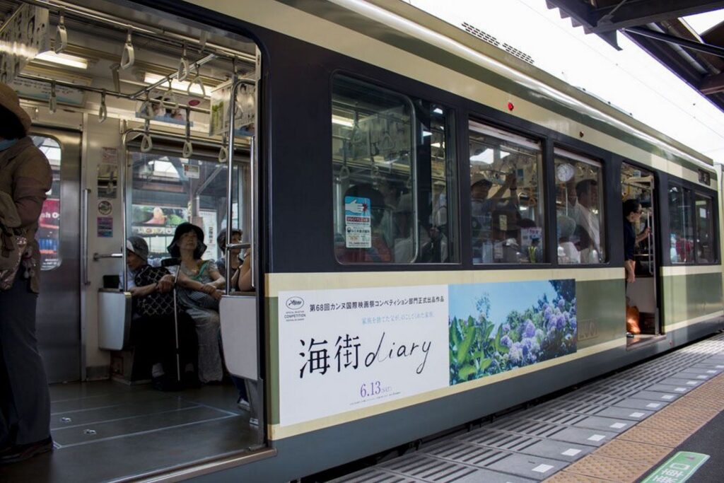 【うみまちダイアリーロケ地】駅のホームに停車する緑とクリーム色の電車側面に海街ダイアリーの広告車内には乗客が見える