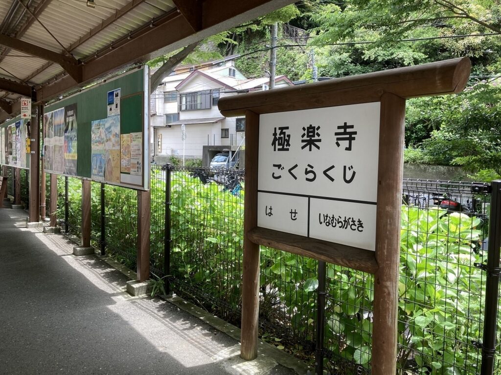 【うみまちダイアリーロケ地】 極楽寺駅の駅名標とホームの風景 緑の植物と掲示板がある