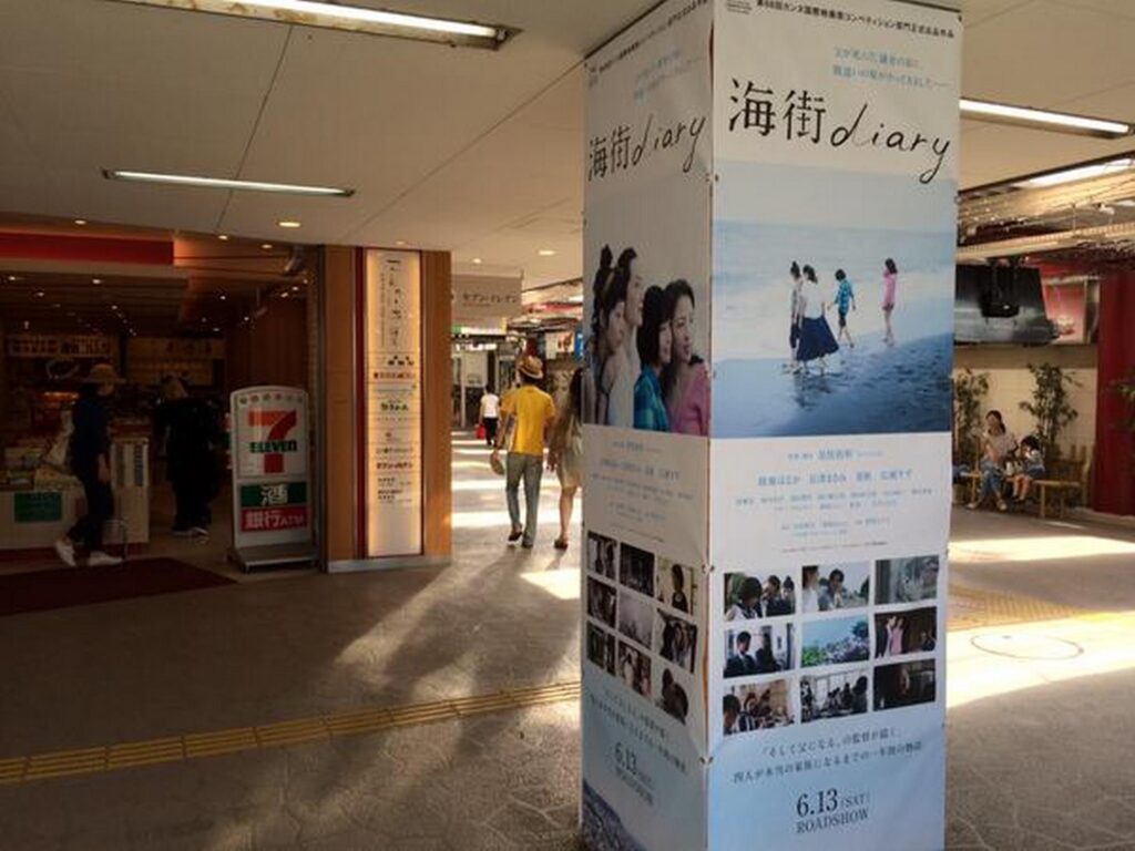 【うみまちダイアリーロケ地】映画海街diaryの大きなポスターがある通路セブンイレブンの店と人々が見える