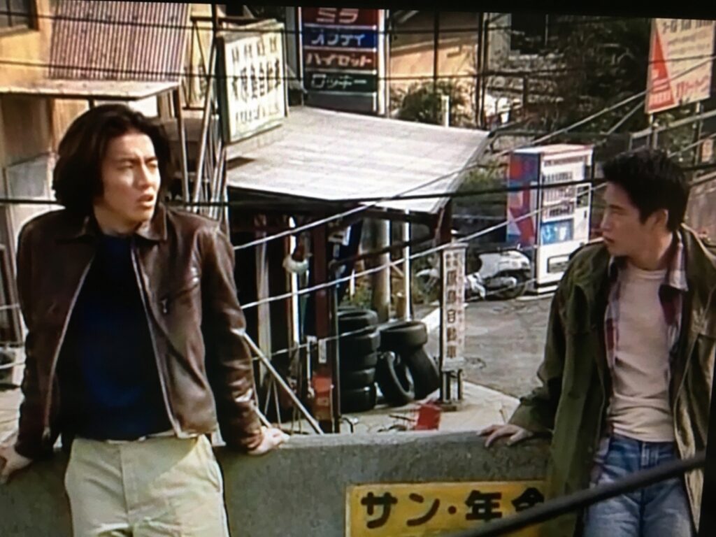 【若者のすべて ロケ地】茶色いレザージャケットの男性とカーキ色のジャケットの男性が古い建物と商店の並ぶ街角に並んで立つ