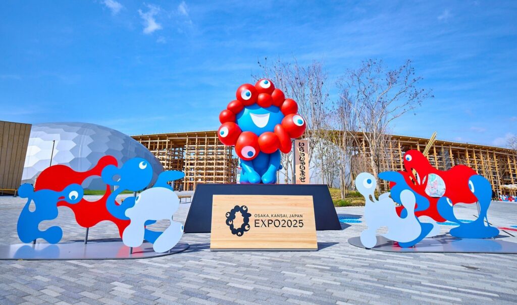 【Expo2025 Osaka】The colorful mascot characters of the Osaka Kansai Expo stand in front of the expo symbol and building with a blue sky background