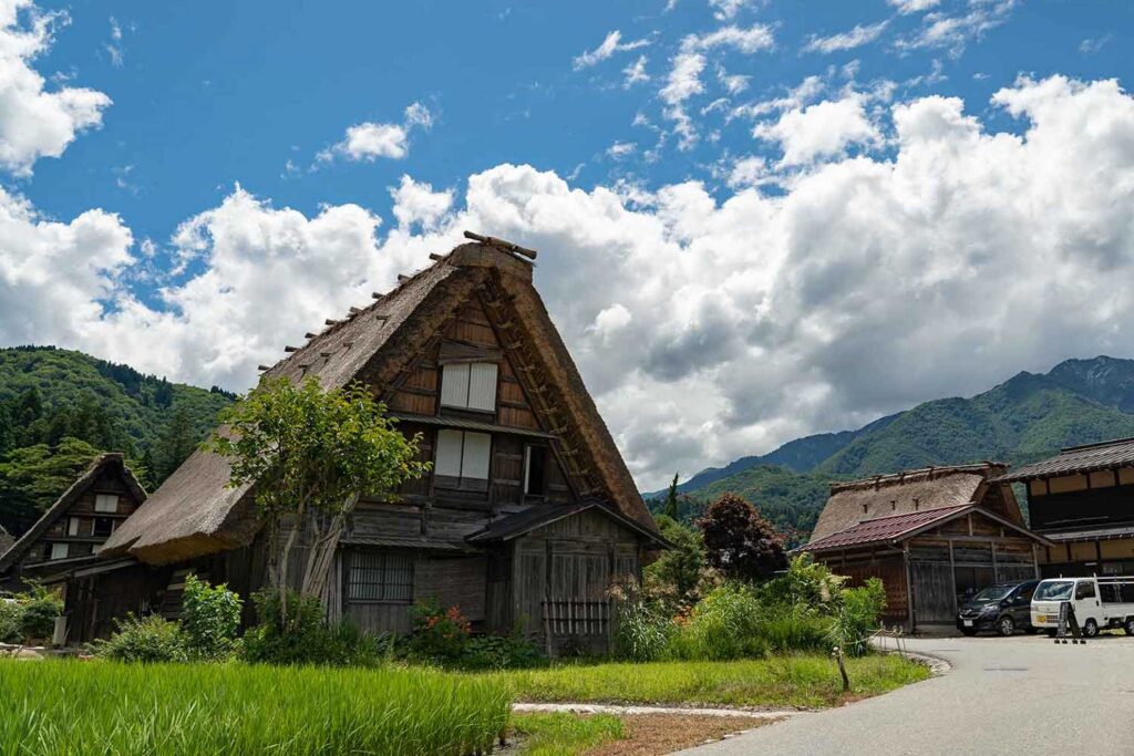 【Shirakawa-go】A traditional Japanese gassho-zukuri house with a thatched roof surrounded by green rice fields and mountains under a cloudy blue sky